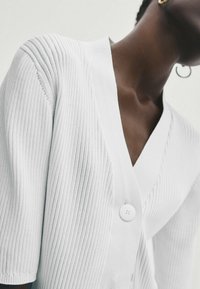 Weißer, gerippter Cardigan mit tiefem V-Ausschnitt, kurzen Ärmeln und einem einzigen weißen Knopfverschluss. Weiche Textur, minimalistisches Design.