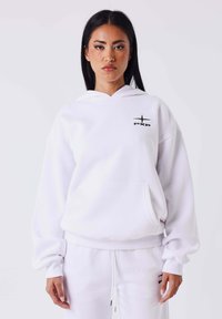 Witte hoodie gemaakt van zachte stof, met een voorzak, geribbelde manchetten en een zwart logo op de borst. Gelijkende broek erbij gedragen.