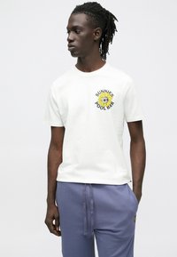 Valkoinen puuvillainen t-paita, jossa on keltainen auringonkukkakuva, jolla on kasvot, ja musta teksti "SUNNIE'S POOL BAR", yhdistettynä sinisiin collegehousuihin.