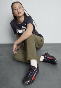 Zapatillas deportivas negras con acentos rojos y púrpuras, parte superior de malla texturizada y suela gruesa. Llevadas con pantalones verde oliva y calcetines blancos.