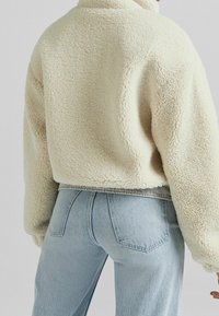 Krämfärgad fleecetröja med texturerad yta, kort design och hög krage, kombinerad med ljusblå jeans.