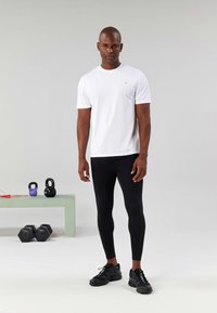 Homem de pé em leggings pretas, t-shirt branca e sapatilhas pretas em um ambiente de ginásio com kettlebells e halteres por perto.