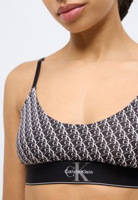Bralette Calvin Klein με ολόλευκο μοτίβο 'CK' σε μαύρο φόντο, ρυθμιζόμενες τιράντες και ελαστική ταινία με το λογότυπο σε ασημί χρώμα.