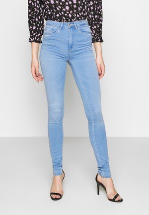 Jeans skinny azul claro de denim, com cintura alta, cinco bolsos e detalhes de costura discretos, combinados com sandálias de salto alto pretas.