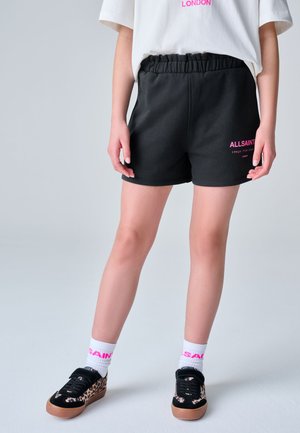 Person iført sorte elastiske taljeshorts med pink tekst, hvide sokker med pink skrift, leopardmønstrede sorte sneakers og en hvid skjorte.