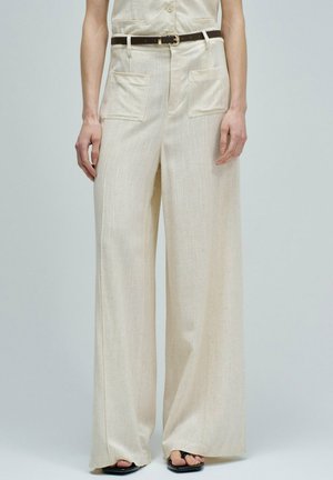 Pantalones beige de pierna ancha con bolsillos delanteros de parche, cinturón negro y sandalias negras abiertas, llevados por una mujer que está de pie frente a un fondo liso.