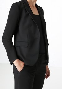 Černé sako na míru s klopovým límcem, zapínáním na jeden knoflík a dvěma předními kapsami; vyrobené z hladké látky.