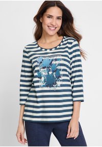 Lächelnde Frau mit langen braunen Haaren, die ein gestreiftes Shirt in Marineblau und Weiß mit einem blauen Blumenmotiv und Marineblaue Hosen trägt.