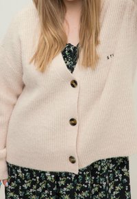 Žena v béžovém cardiganovém svetru s knoflíky, který má přes černé květinové šaty, a s dlouhými blond vlasy, které částečně zakrývají výstřih.