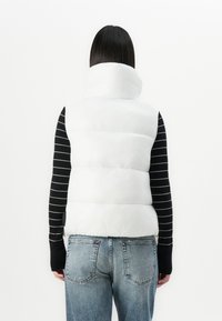 Vrouw met steil zwart haar, die een witte gewatteerde vest, een zwart-wit gestreept langemouwen shirt en vervaagde blauwe jeans draagt, gezien van achteren.