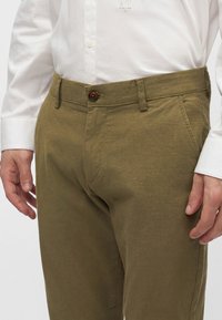 Primo piano di una persona che indossa pantaloni verde oliva con chiusura a bottoni e una camicia bianca a maniche lunghe infilata dentro.