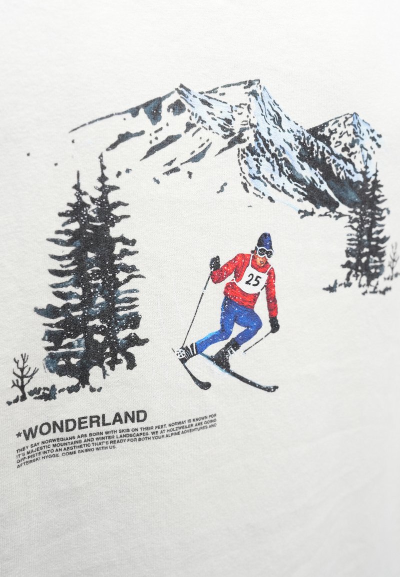 Maglietta bianca con uno sciatore in abbigliamento rosso e blu su uno sfondo di montagne innevate, con il testo nero "WONDERLAND" sotto.
