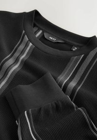 Black sweater with ribbed texture, featuring silver-striped accents across the front and at the cuffs. Label reads "NEXT," size M.