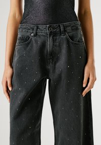 Melna denima bikses ar brīvu piegriezumu, rotātas ar metāliskiem kniedēm un klasisku piecām kabatām. Šaurinātas kājas ar redzamām šuvēm.