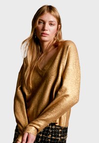 Gul, glittrig tröja med V-ringning, lång ärm och avslappnad passform. Texturen reflekterar ljus och kompletterar en mönstrad kjol.