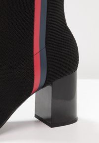 Botín de punto negro con tacón ancho negro, acentuado con franjas laterales rojas y azules en el material superior.