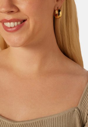 Gold hoop earrings with a polished finish, featuring a curved design. Worn by a model with light hair and a neutral-toned top.