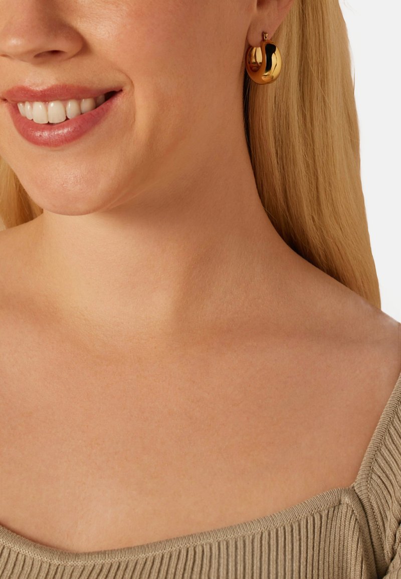 Gouden creolen met een gepolijste afwerking, met een gebogen ontwerp. Gedragen door een model met licht haar en een top in neutrale tinten.
