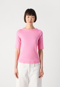Vrouw met kort donker haar, gekleed in een roze top met korte mouwen en hoog opgesneden witte broek, staand tegen een eenvoudige witte achtergrond.