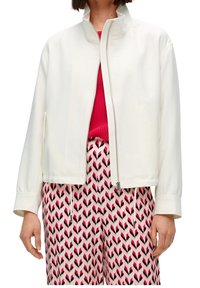 Giacca bianca con zip, colletto alto e maniche lunghe, abbinata a pantaloni a righe rosa e neri con design a zigzag.