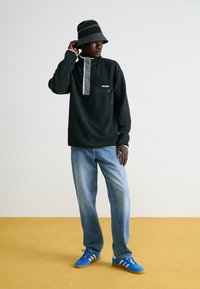 Črn fleec pulover s sivo zapenjanjem na ovratniku, sprednjim žepom, svetlo modrih jeans hlačah, svetlo modrih in sivih supergah, ter črno-belem črtastem klobuku.