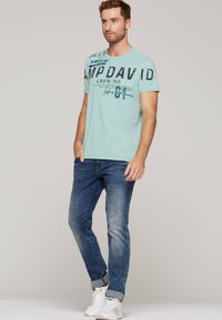 Jasnoniebieska koszulka z odważnym czarnym napisem i graficznymi wzorami, w połączeniu z ciemnoniebieskimi dżinsami i białymi trampkami. Prosty, codzienny strój.