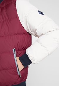 Bordeaux og hvid pufferjakke med en lynlåslomme, ribbede manchetter og et logo-patch på skulderen; har et quiltet design og glat tekstur.