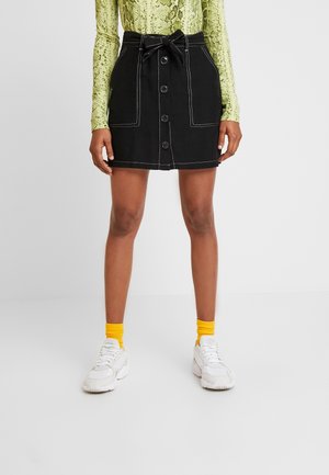 Vrouw in een zwarte rok met knopen aan de voorkant en een gestrikt tailleband, gele sokken, witte sneakers en een groene top met slangenprint met lange mouwen.
