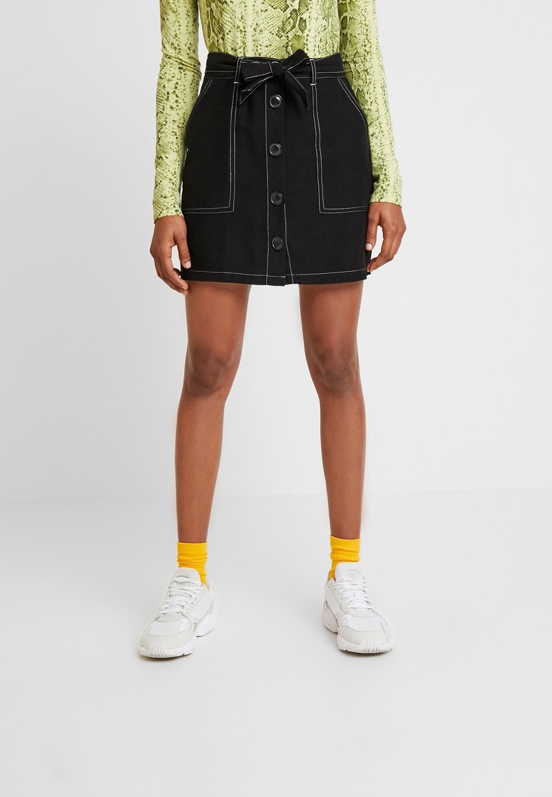 Žena oblečená v černé sukni s knoflíky vpředu a zavázaným páskem, žlutých ponožkách, bílých teniskách a dlouhém zeleném topu s potiskem hada.