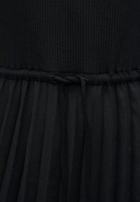 Μαύρο φόρεμα με ριμπ υφαντό πάνω μέρος και ρέον, πιετέ φούστα. Διαθέτει ζωνάκι με κορδόνι για ρύθμιση. Απαλή υφή με ματ φινίρισμα.