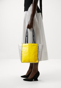 Borsa tote trasparente con davanti giallo, rifiniture blu e manici neri, riportante la scritta "Nørgaard" in bianco. Forma strutturata. Superficie liscia.