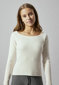 Weißer, gerippter Pullover mit langen Ärmeln, weitem Ausschnitt und figurbetontem Schnitt, kombiniert mit grauen Jogginghosen mit Kordelzug, vor einem neutralen Hintergrund.