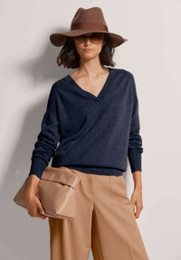 Frau mit einem breiten braunen Hut, dunklen Sonnenbrillen, einem marineblauen V-Ausschnitt-Pullover, beigen weit geschnittenen Hosen und einer großen beigen Clutch.