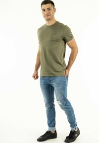 T-shirt kaki a maniche corte con scollo rotondo, abbinato a jeans strappati azzurri chiari e sneaker nere con accenti di design minimi.