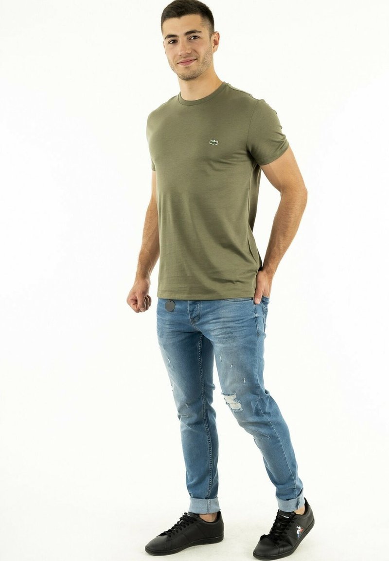 Olivgrünes kurzärmliges T-Shirt mit Rundhalsausschnitt, kombiniert mit hellblauen, zerrissenen Jeans und schwarzen Sneakers mit minimalen Designelementen.