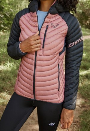 Osoba na zewnątrz w różowo-czarnej pikowanej kurtce i czarnych legginsach, z jedną ręką poprawiającą zamek kurtki.