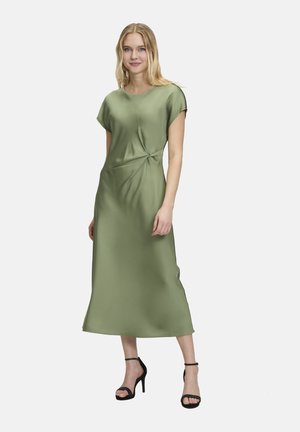Satin, grünes Midi-Kleid mit kurzen Ärmeln, das ein geknotetes Detail an der Taille und eine glatte Textur aufweist, kombiniert mit schwarzen Absätzen.