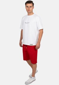 Witte katoenen T-shirt met een zwart logo, gecombineerd met rode katoenen shorts met een elastische tailleband en zak. Witte sneakers maken de look compleet.