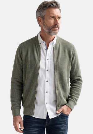 Man with gray hair and beard wearing an olive green textured zip-up jacket over a white button-up shirt and dark jeans, looking to the side.