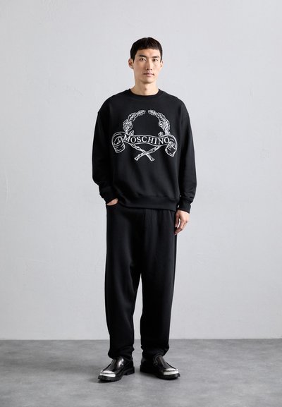 Junger Mann, der drinnen steht und einen schwarzen Moschino-Pullover, lockere schwarze Hosen und schwarze Lederschuhe trägt, vor einer schlichten grauen Wand.
