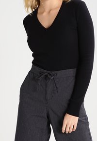Femme portant un haut noir à manches longues et en V, associé à un pantalon taille haute à rayures gris foncé avec une ceinture nouée.