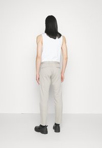 Person med långt svart hår som bär en vit ärmlös topp, ljusbeiga byxor och svarta skor, står vänd mot en vit vägg.