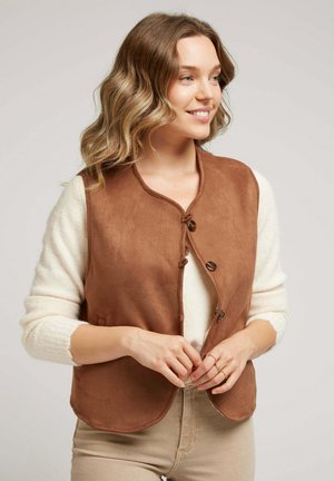 Femme aux cheveux ondulés souriante, portant un pull blanc et boutonnant un gilet sans manches marron sur un pantalon beige, sur fond neutre.