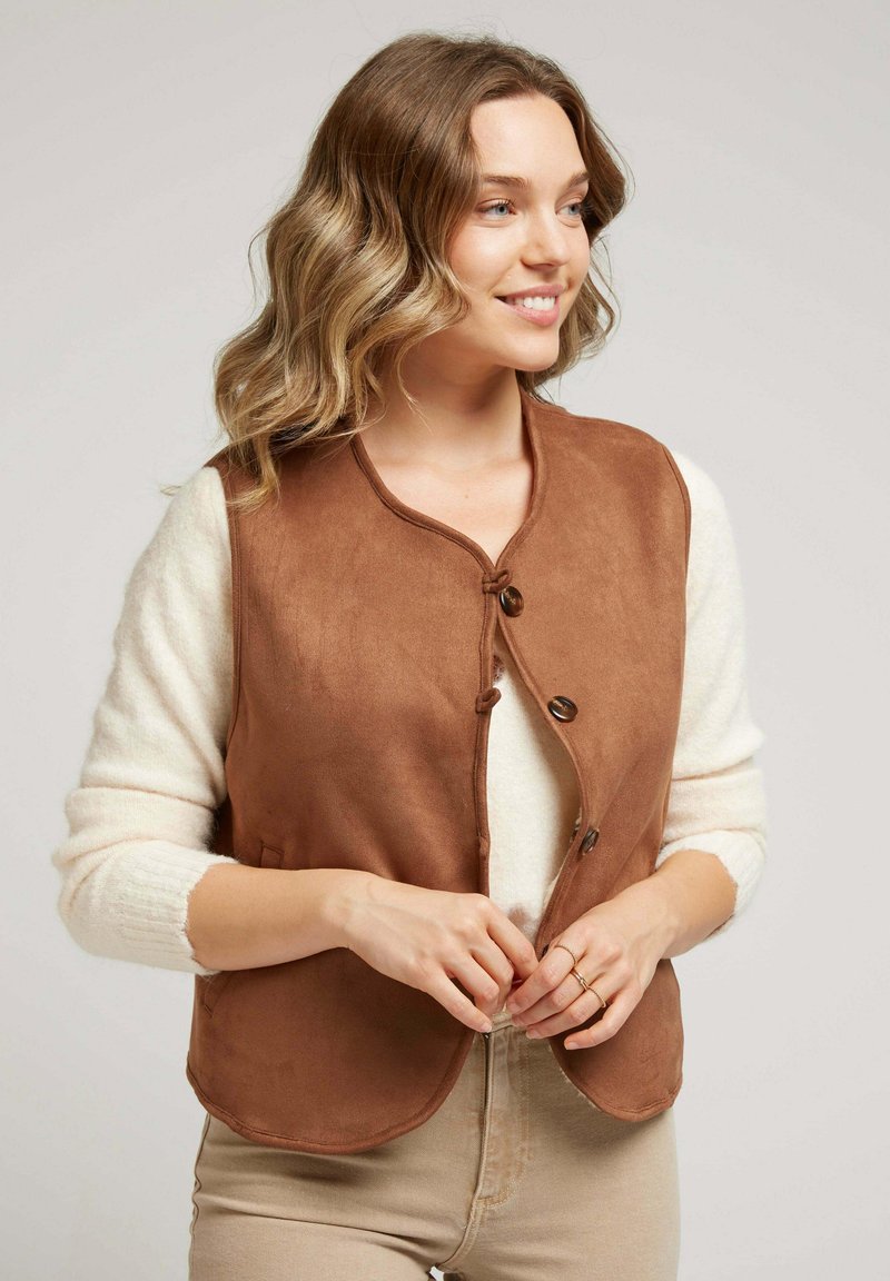 Femme aux cheveux ondulés souriante, portant un pull blanc et boutonnant un gilet sans manches marron sur un pantalon beige, sur fond neutre.