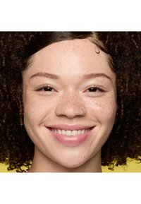 Jeune personne aux cheveux bouclés, taches de rousseur, anneau au nez et boucles d'oreilles souriant sur un fond jaune.
