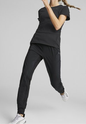 Woman in black athletic wear and white sneakers mid-jump against a neutral gray background, hair in a braid, with arms raised.