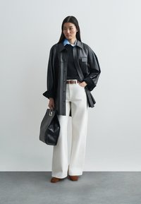 Black leather oversized shirt, navy sweater, white wide-leg trousers, brown heeled shoes, and a black tote bag with a silver accent.