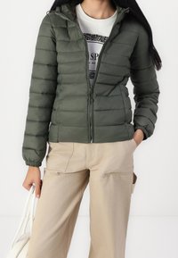 Oliwkowa kurtka puchowa z pikowanym wzorem, na zamek błyskawiczny, z elastycznymi mankietami i kapturem. W zestawie beżowe spodnie cargo i lekka bluzka.
