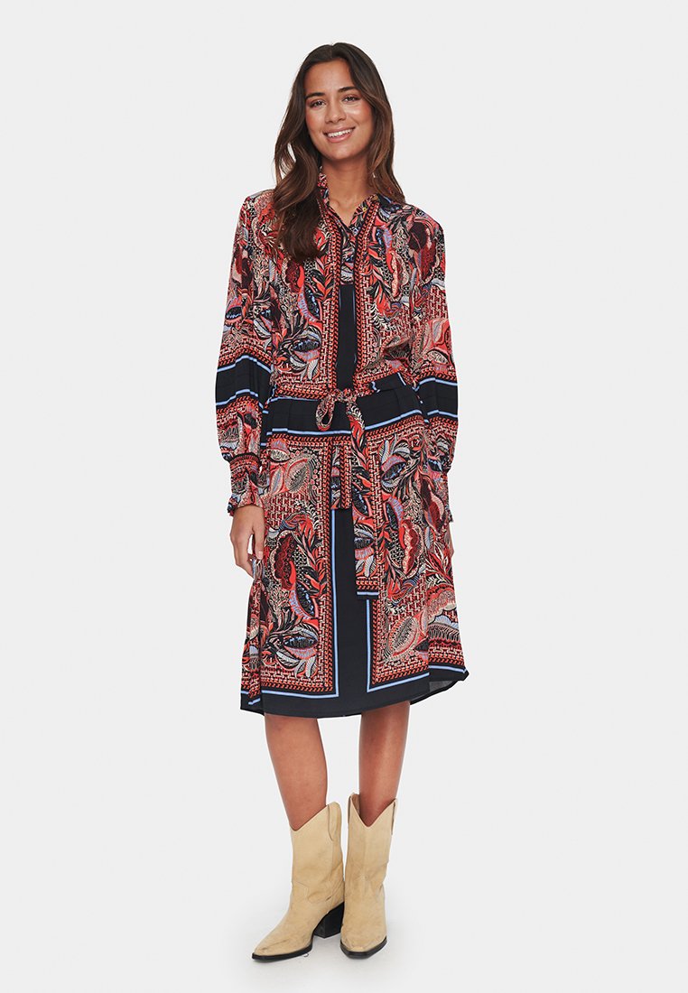 Sukienka z długim rękawem w kolorze granatowym z czerwonym, niebieskim i białym wzorem paisley, z wycięciem w talii i paskiem, oraz długością do kolan. Zestawiona z beżowymi kowbojkami.