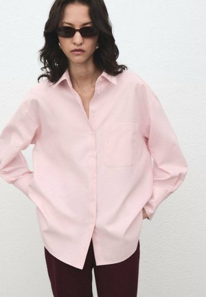 Light pink oversized button-up shirt with collar, single chest pocket, long sleeves, and smooth fabric texture. Paired with dark burgundy pants.